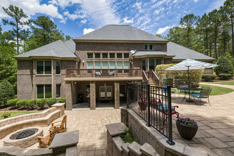 The back of a two-storey house with a raised deck, a paved patio with a sunken fire pit and Adirondack chairs, bordered by lawn and pine trees.